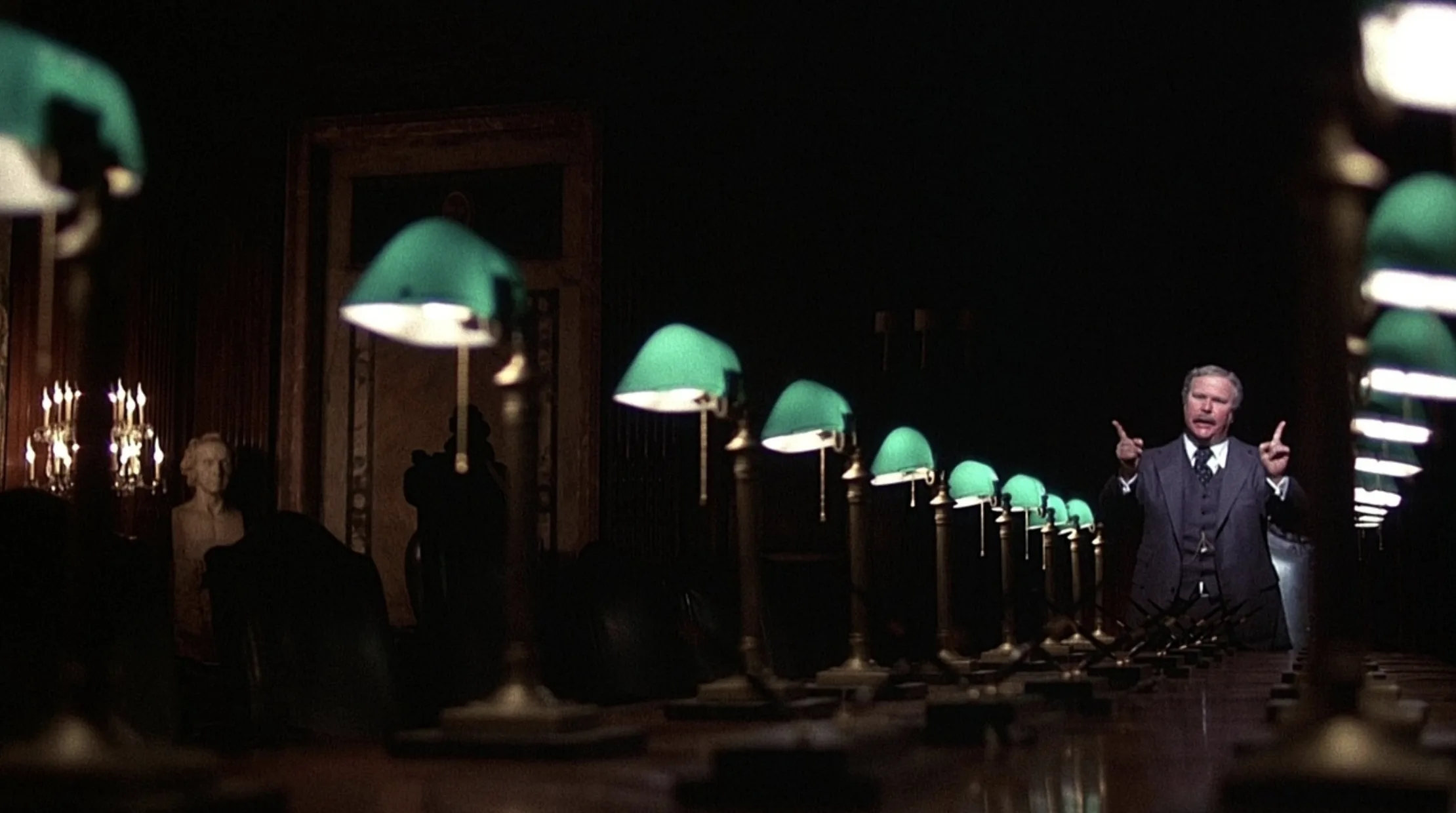 A still from the 1976 film Network: a lone figure stands at the far end of a long, dark boardroom table lined with green banker’s lamps, arms raised.