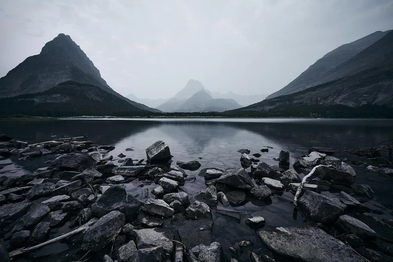 A still mountain lake ringed by peaks, with a rocky foreground shoreline under an overcast sky.
